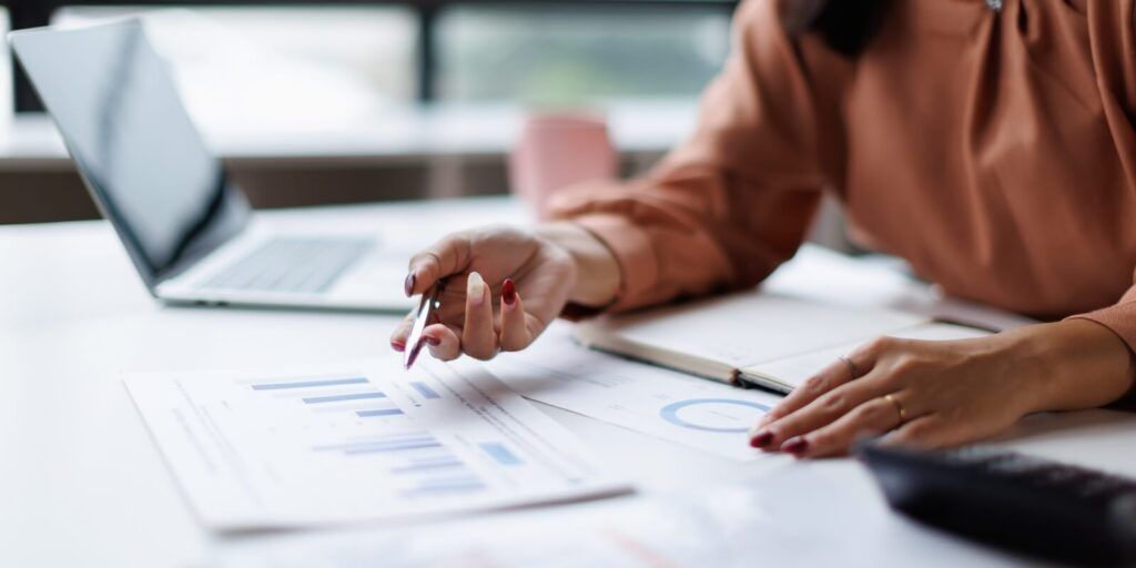 professional woman analyzing financial data and business reports, using a pen to point at charts while working on a laptop at an office desk