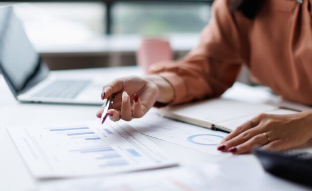 professional woman analyzing financial data and business reports, using a pen to point at charts while working on a laptop at an office desk