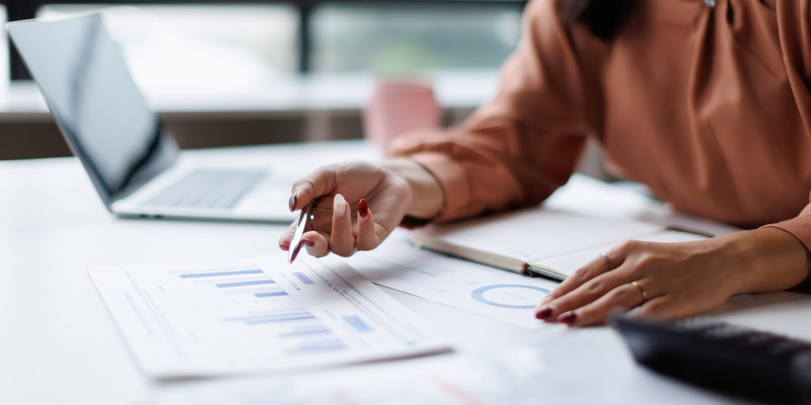 professional woman analyzing financial data and business reports, using a pen to point at charts while working on a laptop at an office desk